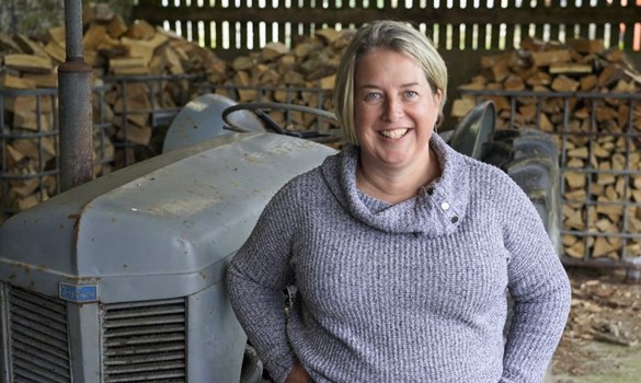 Woman smiling at camera stood in front of an old grey tractor and large stack of logs